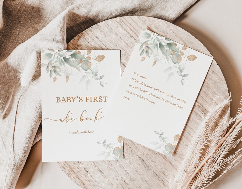 May include: A white card with gold lettering that says "BABY'S FIRST abc book - made with love -" with a watercolor illustration of green leaves and gold accents.  The card is on a wooden surface with a white cloth in the background.