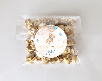 May include: A clear plastic bag filled with caramel popcorn with a round sticker attached to the front. The sticker features a cartoon brown bear holding blue and pink balloons, with clouds and stars in the background. The text on the sticker reads "Ready to pop!"