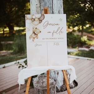May include: A gender reveal sign featuring a brown teddy bear holding balloons. The sign reads "Gar&ccedil;on ou Fille? Qu'en pensez vous?" with a space to write "Gar&ccedil;on" or "Fille".