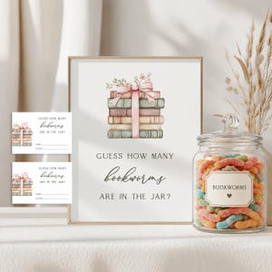 May include: A game set featuring a vertical sign and two small cards with the text GUESS HOW MANY bookworms ARE IN THE JAR? and a watercolor illustration of stacked books tied with a pink ribbon, alongside a glass jar filled with gummy worms.