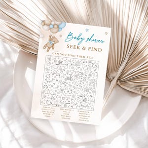 Puede incluir: Una tarjeta de juego de b&uacute;squeda para baby shower con una ilustraci&oacute;n de oso de peluche y el texto "Baby shower SEEK & FIND CAN YOU FIND THEM ALL?". La tarjeta presenta una variedad de peque&ntilde;as ilustraciones en blanco y negro de art&iacute;culos para beb&eacute;s. La tarjeta est&aacute; sobre un plato blanco.