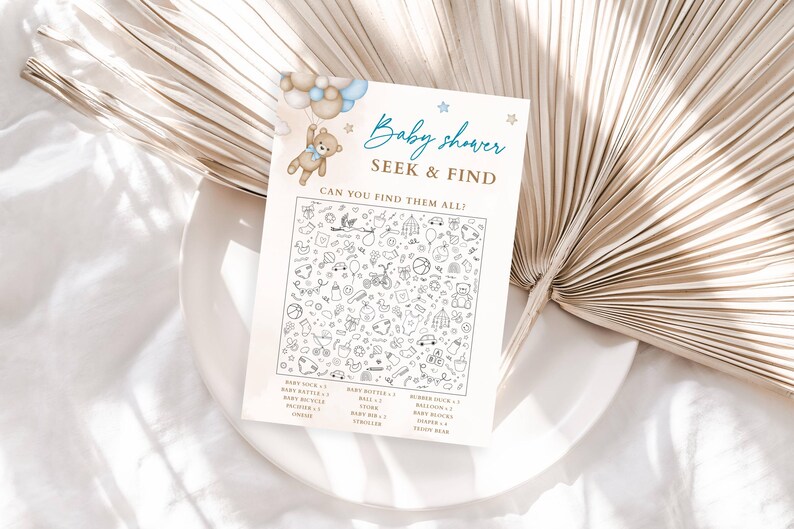 Puede incluir: Una tarjeta de juego de b&uacute;squeda para baby shower con una ilustraci&oacute;n de oso de peluche y el texto "Baby shower SEEK & FIND CAN YOU FIND THEM ALL?". La tarjeta presenta una variedad de peque&ntilde;as ilustraciones en blanco y negro de art&iacute;culos para beb&eacute;s. La tarjeta est&aacute; sobre un plato blanco.