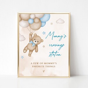 Puede incluir: Impresi&oacute;n enmarcada con borde dorado. Un oso de peluche con lazo azul sostiene globos marrones, blancos y azules. El texto dice "Mommy's cravings station" y "A few of mommy's favorite things". El fondo es beige.