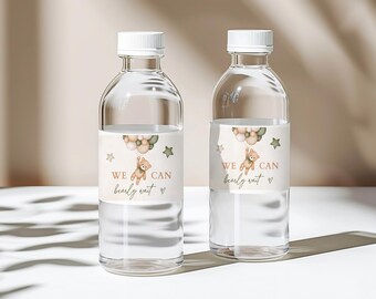 Op de afbeelding: Twee heldere glazen waterflessen met witte etiketten. De etiketten tonen een cartoonbeer die ballonnen vasthoudt en de tekst "We can barely wait".