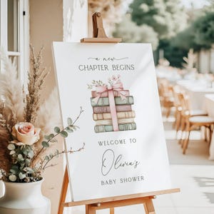 May include: A white sign on a wooden easel with the text "a new chapter begins" and "Welcome to Olivia's Baby Shower." The sign features a stack of books tied with a pink ribbon and floral accents. A vase with flowers is in the foreground.