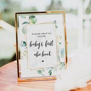 May include: Gold framed sign with a white background and green watercolor leaves. The sign says "Please help us color baby's first abc book".