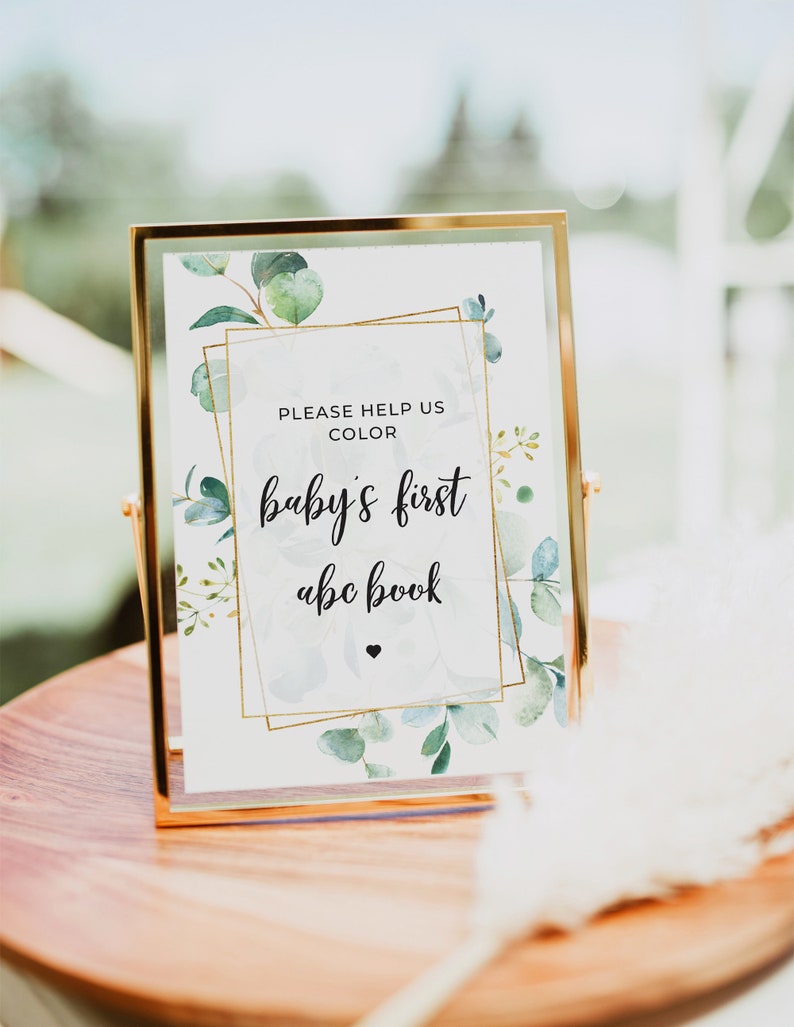 May include: Gold framed sign with a white background and green watercolor leaves. The sign says "Please help us color baby's first abc book".