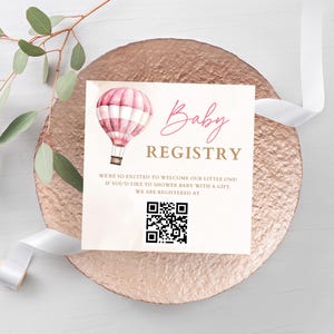 Może przedstawiać: Karta z ogłoszeniem rejestracji dziecka z akwarelową ilustracją balonu na ogrzane powietrze. Karta znajduje się na powierzchni o fakturze różowego złota z napisem "Baby Registry". Dołączony jest kod QR.
