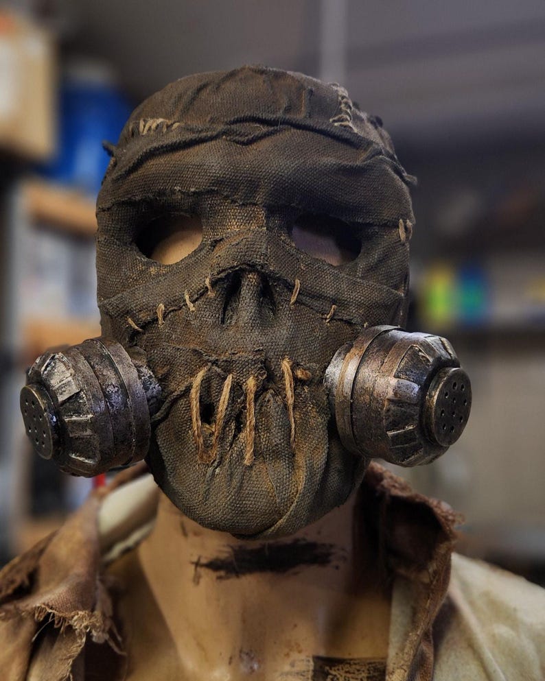 Może przedstawiać: Brązowa maska z juty z przeszytymi detalami i otworami na oczy. Maska ma przeszyte usta i dwa metalowe pojemniki w stylu przemysłowym po obu stronach. Maska jest częścią kostiumu.