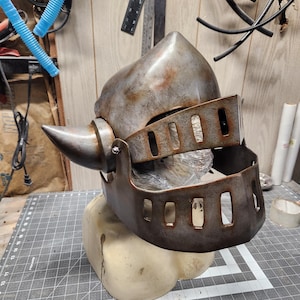 Könnte beinhalten: Ein verwitterter Ritterhelm aus Metall mit einem aufklappbaren Visier und einer spitzen Spitze. Der Helm hat ein abgenutztes, gealtertes Aussehen mit rostfarbenen Akzenten. Er ruht auf einer beigen Form, die einen Ausstellungsständer andeutet. Das Visier hat rechteckige Aussparungen.