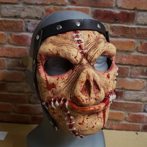 Puede incluir: Una máscara espantosa con un diseño cosido y salpicaduras de sangre. La máscara presenta una parte superior gris en forma de casco con una correa negra y tachuelas plateadas. La cara es de tono carnoso con acentos rojos.