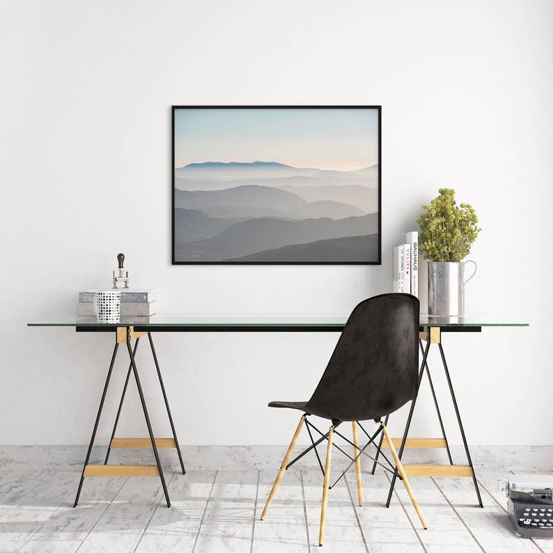 Op de afbeelding: Een zwart ingelijste foto van een bergketen met een wazige, blauwe lucht. De foto hangt aan een witte muur boven een glazen bureau met een zwarte stoel. Het bureau wordt ondersteund door twee gouden en zwarte bokpoten.