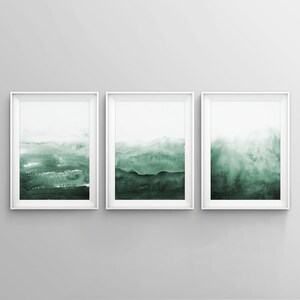 May include: Three framed watercolor paintings with a green and white abstract landscape design. The paintings are in white frames and are hung on a white wall.