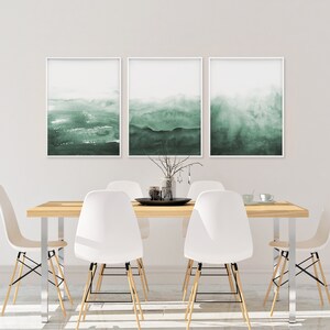May include: Three framed watercolor paintings with a green and white color scheme depicting abstract landscapes. The paintings are hanging on a white wall above a wooden dining table with white chairs.