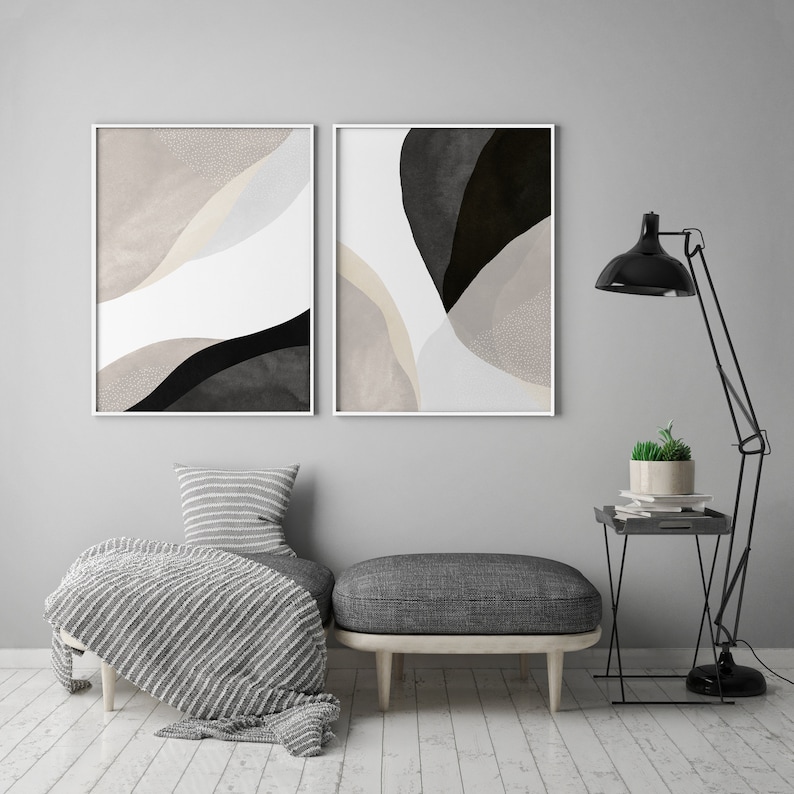 May include: Two abstract art prints in white frames with black and beige shapes on a white background. The prints are hanging on a gray wall above a gray ottoman with a white and gray striped throw blanket.