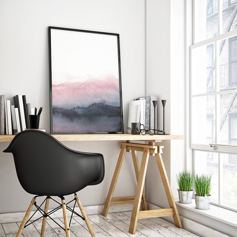 May include: A black framed watercolour print of a pink and blue landscape hangs above a wooden desk with a black chair. The desk is supported by two wooden sawhorses. There are two potted plants on the windowsill.