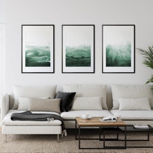 May include: Three framed abstract watercolor paintings in shades of green, hanging on a white wall above a white sectional sofa with black throw pillows and a wooden coffee table.