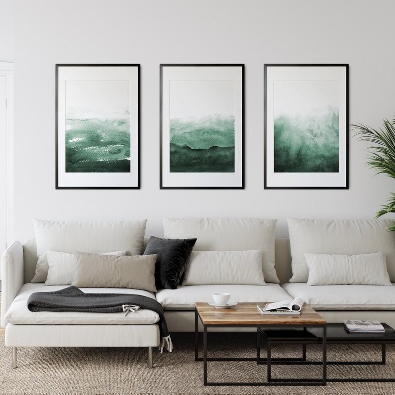 May include: Three framed abstract watercolor paintings in shades of green, hanging on a white wall above a white sectional sofa with black throw pillows and a wooden coffee table.