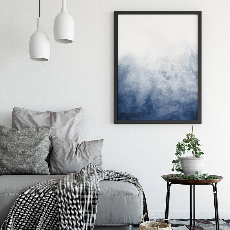 Op de afbeelding: Een blauw aquarel schilderij in een zwart frame hangt aan een witte muur boven een grijze bank met kussens en een deken. Een kleine zwarte bijzettafel met een plant in een pot staat in de hoek van de kamer.