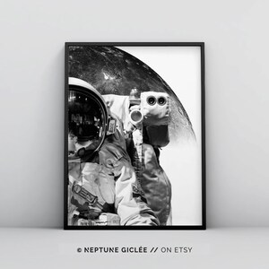 May include: Black and white photograph of an astronaut in a spacesuit standing in front of a large, round moon. The astronaut's helmet reflects the moon's surface. The image is framed in a black frame.
