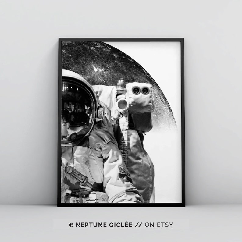 May include: Black and white photograph of an astronaut in a spacesuit standing in front of a large, round moon. The astronaut's helmet reflects the moon's surface. The image is framed in a black frame.