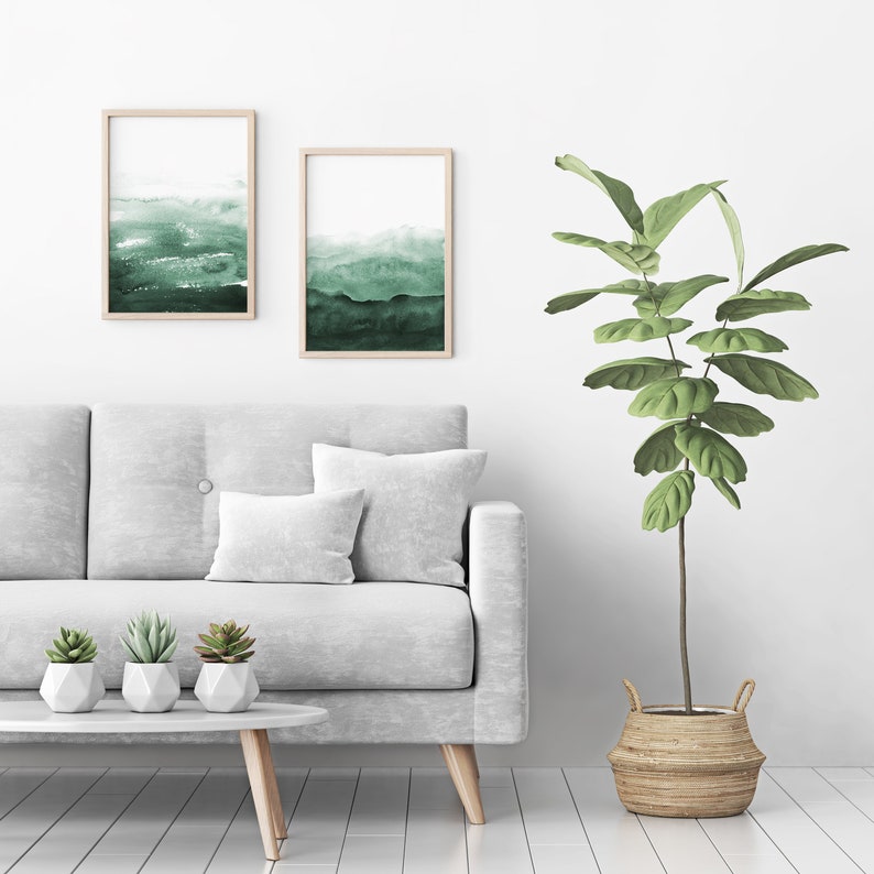 May include: A white sofa with two throw pillows and three small potted succulents sits in front of a white wall with two framed watercolor paintings of green landscapes. A large potted plant with green leaves stands to the right of the sofa.
