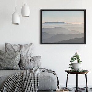 Op de afbeelding: Een zwart ingelijste print van een mistig berglandschap met een lichtblauwe lucht. De print hangt aan een witte muur boven een grijze bank met kussens en een zwart-witte deken.