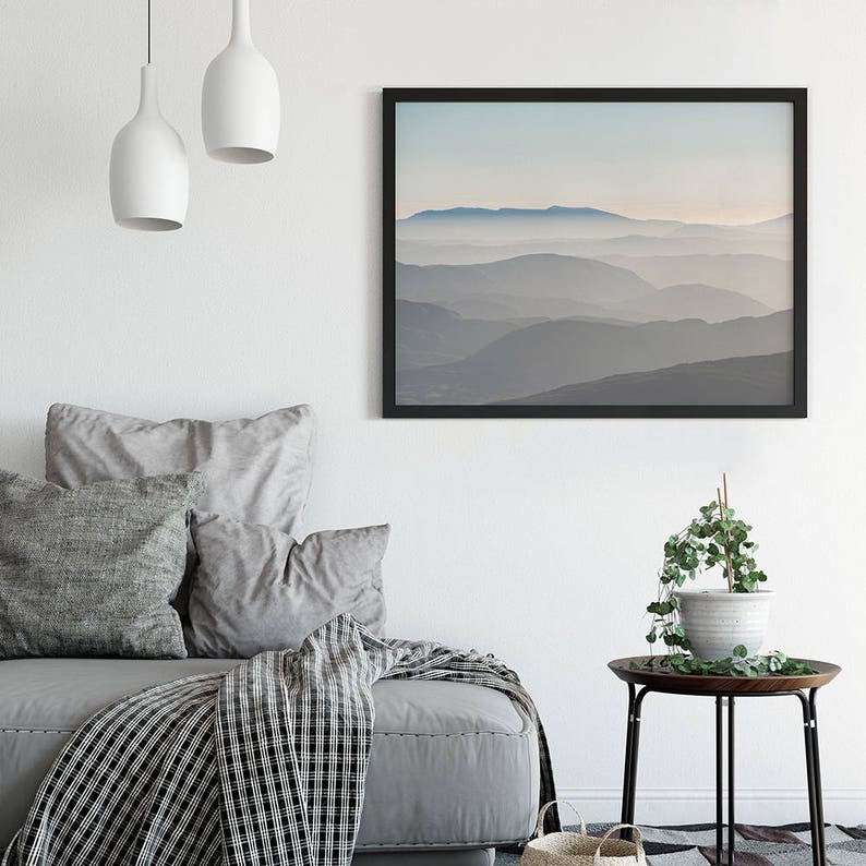 Op de afbeelding: Een zwart ingelijste print van een mistig berglandschap met een lichtblauwe lucht. De print hangt aan een witte muur boven een grijze bank met kussens en een zwart-witte deken.