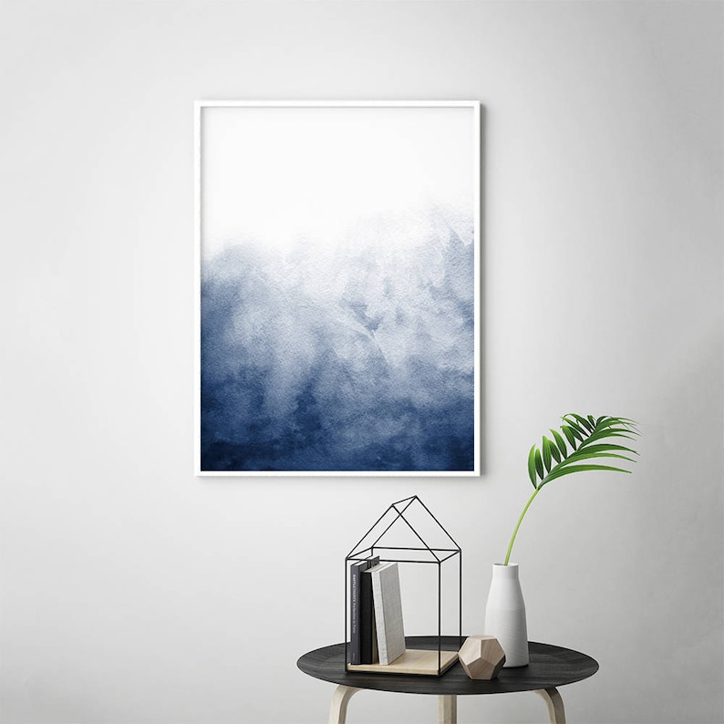 Op de afbeelding: Een ingelijst abstract aquarel schilderij met een blauwe en witte kleurverloop. Het schilderij is in een wit frame en hangt aan een witte muur.