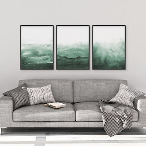 May include: Three framed abstract watercolor paintings in shades of green, hanging on a white wall above a gray couch with pillows and a throw blanket.