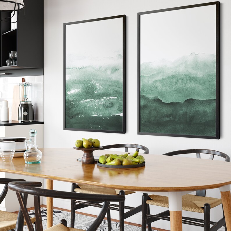 May include: Two framed abstract watercolor paintings in shades of green, hanging on a white wall above a wooden dining table with chairs. The table is set with a bowl of pears.