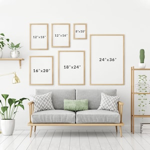 May include: A white wall with a couch and five framed pictures of different sizes. The pictures are all in wooden frames and have measurements written on them in inches. The couch is gray with two pillows and is sitting on a wooden floor.