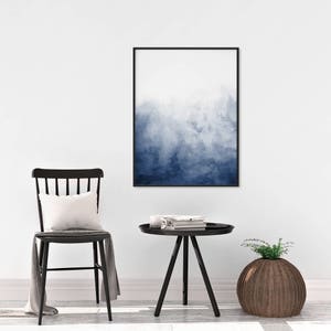 Op de afbeelding: Een zwart ingelijst afdruk van een blauwe aquarel hangt aan een witte muur boven een zwarte bijzettafel met een zwarte stoel en een geweven mand.