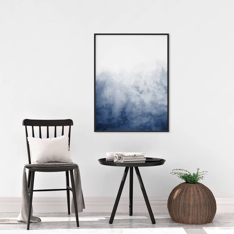Op de afbeelding: Een zwart ingelijst afdruk van een blauwe aquarel hangt aan een witte muur boven een zwarte bijzettafel met een zwarte stoel en een geweven mand.