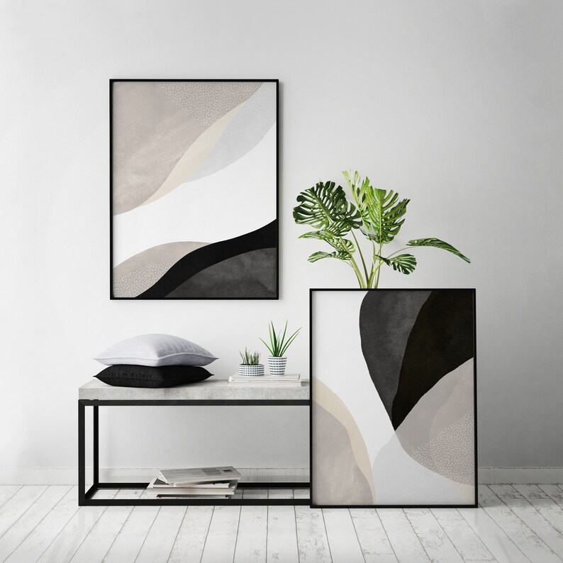 May include: Two black framed abstract art prints with beige, white, and black colors. The prints are hanging on a white wall above a black metal table with a concrete top. A small potted plant is on the table in front of the prints.