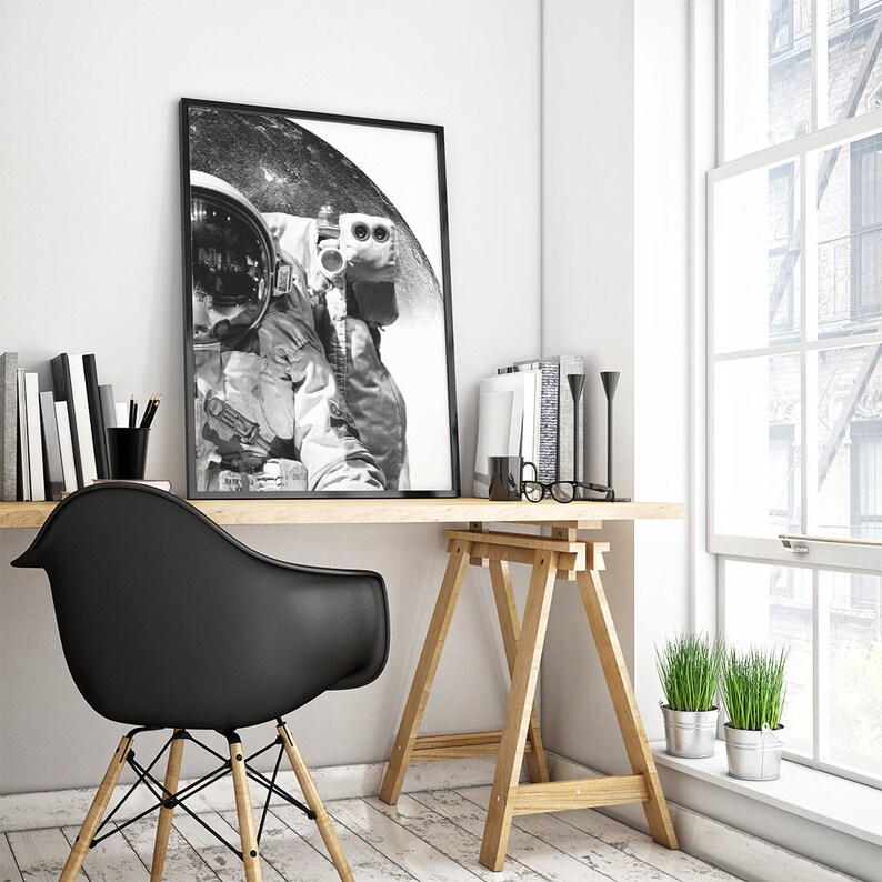 May include: A black and white photograph of an astronaut in a spacesuit, framed in a black frame, hanging on a white wall above a wooden desk with a black chair. The desk is also decorated with books, pencils, and a pair of glasses.