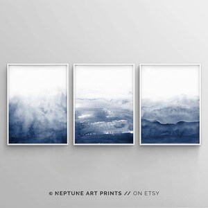 Op de afbeelding: Set van drie abstracte aquarelprints in tinten blauw. De prints zijn ingelijst in witte lijsten en tonen een verloop van lichtblauw naar donkerblauw.