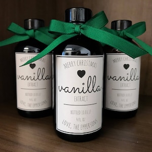 May include: Three glass bottles of vanilla extract with green ribbon bows. The labels are white with black text and a heart. The center bottle reads "Merry Christmas vanilla extract" and "Bottled 10.03.17 4 fl oz Love, The Eppersons".