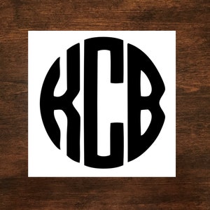 May include: A black monogram design featuring the letters KCB in a bold, rounded font, set within a circular shape. The design is printed on a white square, set against a dark wood grain background.