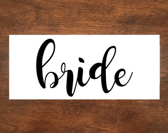 May include: White sign with the word "bride" written in black cursive script.
