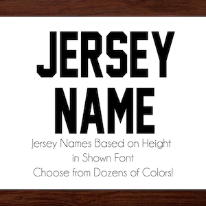 May include: A white rectangular sign with the words "JERSEY NAME" in bold, black block letters. Below, smaller text reads "Jersey Names Based on Height in Shown Font. Choose from Dozens of Colors!" The background is a dark wood.