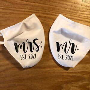 Pu&ograve; includere: Due maschere di stoffa bianche con testo nero. Una maschera dice "mrs. EST. 2021" e l'altra dice "mr. EST. 2021".