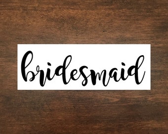 May include: A white rectangular sign with the word "bridesmaid" in black cursive script. The sign is set against a dark brown wooden background.