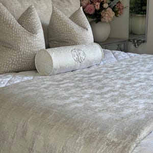 Pode incluir: Uma cama com padrão branco e cinza com um travesseiro com padrão branco e cinza e um travesseiro cilíndrico branco com um monograma. A cama é feita com uma colcha com padrão branco e cinza.