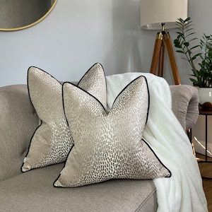 May include: Two beige throw pillows with a black trim and a subtle abstract pattern. The pillows are on a light brown couch with a white throw blanket.