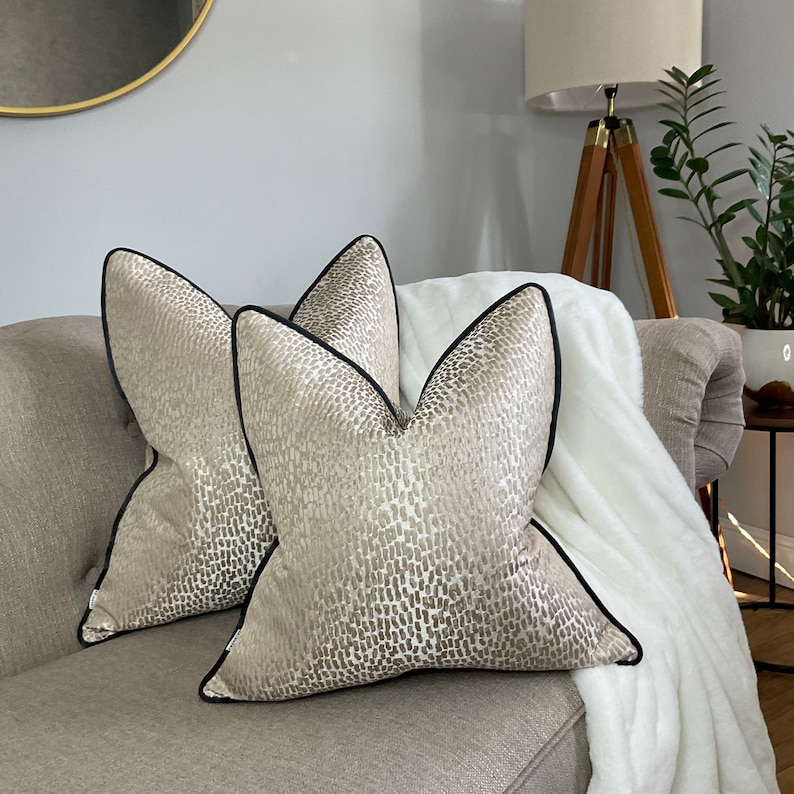 May include: Two beige throw pillows with a black trim and a subtle abstract pattern. The pillows are on a light brown couch with a white throw blanket.