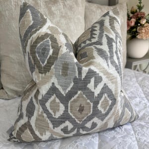 Puede incluir: Una almohada decorativa con un patrón geométrico en tonos grises, marrones y blancos. La almohada está hecha de una tela suave y texturizada.