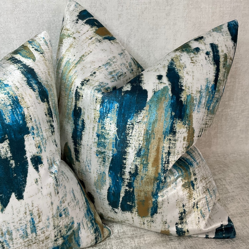 May include: Four decorative throw pillows with a white background and a bold abstract design. The pattern features strokes of teal, blue, and gold, creating a textured, artistic look. The pillows are arranged in a layered composition.