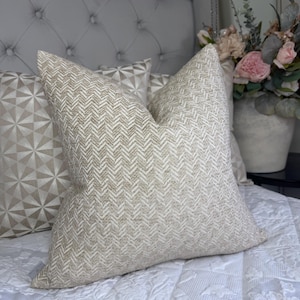 May include: A square throw pillow featuring a textured beige and white herringbone pattern. The pillow is constructed from a woven fabric with a consistent zigzag design across the entire surface, showcasing a neutral color palette and a structured shape.