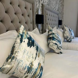 May include: A bedroom scene featuring two upholstered headboards with button tufting. White bedding is topped with decorative pillows in shades of blue, teal, and gold. Black lamps are mounted on the wall between the headboards. The wallpaper has a gray and white pattern.